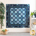 A blue quilt with a diamond pattern hangs on a wall, surrounded by greenery and a shelf with decor.