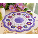 A round floral table mat with a scalloped edge, featuring purple, pink, and blue flowers arranged around a central circle. The mat is placed on a wooden table beside scissors, spools of thread, and decorative buttons, with purple flowers in a vase in the background.