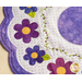 A close-up of a fabric design featuring colorful flowers in shades of purple and blue with yellow centers, set against a white background dotted with small purple circles. The design is framed by a scalloped edge.