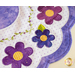 A close-up image of a fabric design featuring purple and blue flowers with yellow centers, set against a background of white polka dots. There are green leaves and decorative stitching in a vine-like pattern. The overall design has a quilted appearance.