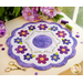 A round purple and white quilted table mat with a scalloped edge, featuring a central purple circle and various colorful flower designs around it. The mat is displayed on a wooden surface alongside scissors, sewing spools, and pink flowers in a jar.