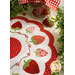 A white countertop with foliage and a houseplant in the background with a white and red strawberry themed table topper with a jar of strawberries in the background