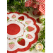 A white countertop with foliage and a houseplant in the background with a white and red strawberry themed table topper with a jar of strawberries in the background