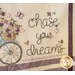 A decorative piece featuring embroidered flowers and butterflies, with the phrase chase your dreams written in a cursive font, accompanied by a bicycle image.