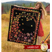 Photo of a blonde woman in an orange dress holding the Autumn Allure quilt in front of her while standing in a field of dry grass with a green mountain in the background and a red wagon at her side.