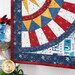Angled close up photo of finished patriotic quilt square on a craft hanger sitting on a brown countertop with a white paneled wall in the background and patriotic decor and plants to one side.