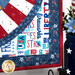 Close up of patriotic quilt square made with red, white, blue, and yellow fabrics with stars and textures in a radiating pattern on a countertop with patriotic decor in the background.