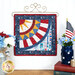 Photo of finished patriotic quilt square on a craft hanger sitting on a brown countertop with a white paneled wall in the background and patriotic decor on either side.