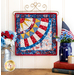 Photo of finished patriotic quilt square on a craft hanger sitting on a brown countertop with a white paneled wall in the background and patriotic decor on either side.