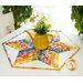 A yellow teapot sits on a colorful hexagonal table runner adorned with floral patterns, accompanied by a small bouquet of flowers in the teapot. The background features plants and natural light, creating a bright and cheerful atmosphere.