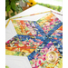 A colorful quilted table runner with a star pattern, featuring vibrant floral and geometric designs in a range of colors, set on a white surface with a blurred background of green leaves and soft sunlight.