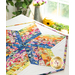 A colorful quilt with a hexagonal pattern featuring vibrant floral fabrics, displayed on a table with a soft background of a window and greenery.