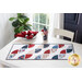 A white table in front of a blue wall with a bright window and green houseplant in the background and a patriotic-themed table runner with a stack of white plates and matching cloth napkins with an American flag centerpiece on the table in the back.