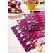 A set of colorful fabric placemats featuring floral designs in shades of purple, pink, and green, arranged neatly on a white table. In the background, there are rolled napkins with a coordinating design and a yellow pitcher with pink flowers.