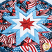 A quilt featuring a star design with red, white, and blue fabric. The pattern includes elements resembling the American flag with wavy stripes and stars arranged in a circular layout.