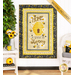 Photo of finished panel quilt hanging on a white paneled wall with a houseplant on one side, sunflower and bee decor, and white wicker furniture with a banner in the top right corner that reads 