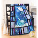 A decorative quilt hangs over a black wooden chair, featuring a wintry scene with a church and snowflakes, framed by colorful patches of blue and purple fabric. The setting is cozy, with light-colored wood floors and a simple, festive backdrop.