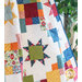 A colorful quilt featuring a patchwork design with stars and various floral and geometric patterns, set against a white background. There is a green leafy plant visible in the background.