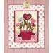 A decorative fabric wall hanging features a pot of flowers with heart-shaped blooms in shades of pink and blue, set against a light floral background and bordered with a red frame.