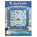 A decorative quilt titled The Joy of Winter featuring various winter-themed appliqués and designs, including snowmen, mittens, trees, and snowflakes, surrounded by borders in shades of blue. The quilt is displayed against a neutral wall with accent furnishings around it.