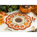 Photo of a scalloped table mat with pumpkins in a ring and embroidered details on a white table with a pair of scissors, threads, and pumpkin decor.