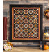 Photo of an orange, brown, and black quilt featuring traditional quilt block patterns hanging on a brown paneled wall with fall decor all around
