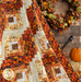 Close up of the log cabin throw quilt made with orange, brown, and cream fabrics draped in the foreground with a dark paneled wall, fall wreath, and pumpkin decor in the background