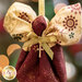 A handcrafted angel ornament made from fabric, featuring a round head and large, decorative fabric wings. The body is draped in burgundy cloth, and a gold ribbon with a small cross is tied around the waist. The background is softly blurred with light bokeh effects.