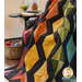 Photo of the Scintillation flannel quilt made with rainbow colored fabrics and black accents draped over a chair in a brightly lit room with a basket of yarn and a table with a lamp, plant, and books in the background