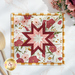 Close up photo of a square folded star hot pad made with pink, white, and red floral fabrics with yellow gingham laying flat on a white countertop with wooden spoons and a bouquet of flowers