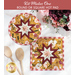 Photo of two folded star hot pads, one round and one square, made with yellow, white, and red floral fabrics laying flat on a white countertop with wooden spoons and a bouquet of flowers