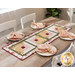 Photo of a table runner made with log cabin blocks and cream, yellow, green, and red floral fabrics laying in the center of a table with light wood and white chairs and four place settings at each seat with matching napkins