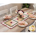 Photo of a table runner made with log cabin blocks and cream, yellow, green, and red floral fabrics laying in the center of a table with a bouquet of flowers in a vase at the center with light wood and white chairs and four place settings at each seat with matching napkins