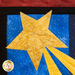 A bright yellow star with rays extending outward, set against a blue background with a starry pattern. The star is outlined by a black border and surrounded by a red quilted area.