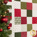 Close up photo of a green, red, and cream Christmas-themed quilt flat against a wall with a decorated Christmas tree in the foreground