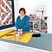 woman using a quilting ruler on a cutting mat to cut fabric next to a quilt