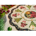 A circular Christmas table mat featuring a festive design with green Christmas trees, candy canes, and red bows arranged around a central image of a golden ornament and poinsettias. The mat has a scalloped edge and is surrounded by holiday decorations, including ornaments and greenery.