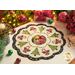 A decorative holiday table centerpiece with a floral and Christmas tree design, featuring green trees, red poinsettias, and candy canes, set on a light background with a scalloped edge. Surrounding the centerpiece are festive decorations including a lit candle and ornaments.