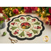 A decorative holiday table centerpiece with a floral and Christmas tree design, featuring green trees, red poinsettias, and candy canes, set on a light background with a scalloped edge. Surrounding the centerpiece are festive decorations including a lit candle and ornaments.