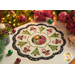 A decorative holiday table centerpiece with a floral and Christmas tree design, featuring green trees, red poinsettias, and candy canes, set on a light background with a scalloped edge. Surrounding the centerpiece are festive decorations including a lit candle and ornaments.