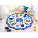 A circular quilted table centerpiece features a blue and white design with various patterns of mittens and a central circular motif. The backdrop includes winter-themed decorations like white snowflakes and small candles.