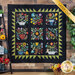 Photo of the finished Folk Art Sampler quilt hanging on a brown paneled wall with a tall shelf on one side decorated in colorful items like a beehive, potted plants, and red flowers, with a basket of flowers and house plant on the other side.