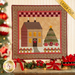 A cream paneled wall with a small wall hanging depicting a house in a snowy field with a Christmas tree in the yard decorated with wrapped gifts. Christmas decor is all around the wall hanging with a decorated tree in the foreground.