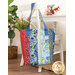 Photo of a large jelly roll tote made with bright floral fabrics filled with flowers hanging from a small white chair with a white table, bouquet, and house plant in the background.