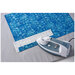 An iron is used to flatten blue fabric on top of a white ruler with dark measurement lines.