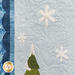 A close-up image of a quilted fabric featuring a light blue background with white snowflakes and a green tree with a snow-covered top. The quilt has textured patterns and is framed by darker blue fabric on the side.