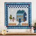 A quilted wall hanging featuring a blue house with a snowman and a tree in a winter scene. The background has a border with a checkered pattern and snowflakes. There is a small wooden fence in front of the house. A banner in the top corner reads LASER CUT.