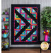 A vibrant quilt featuring a geometric pattern in bright colors, predominantly arranged in a log cabin style, set against a black background. The quilt is displayed on a wall in a room with wooden flooring and decor items, including shelves with books and plants.