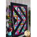 A vibrant quilt is displayed on a wall, featuring a log cabin pattern with bright colors and a black background. It is surrounded by a decorative shelf with potted plants and colorful items.