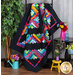 A colorful quilt hangs on a display rack, featuring a zigzag pattern in vibrant shades of pink, blue, green, and yellow against a black background. Nearby, there are potted plants, a small yellow chair, and bouquets in decorative vases.